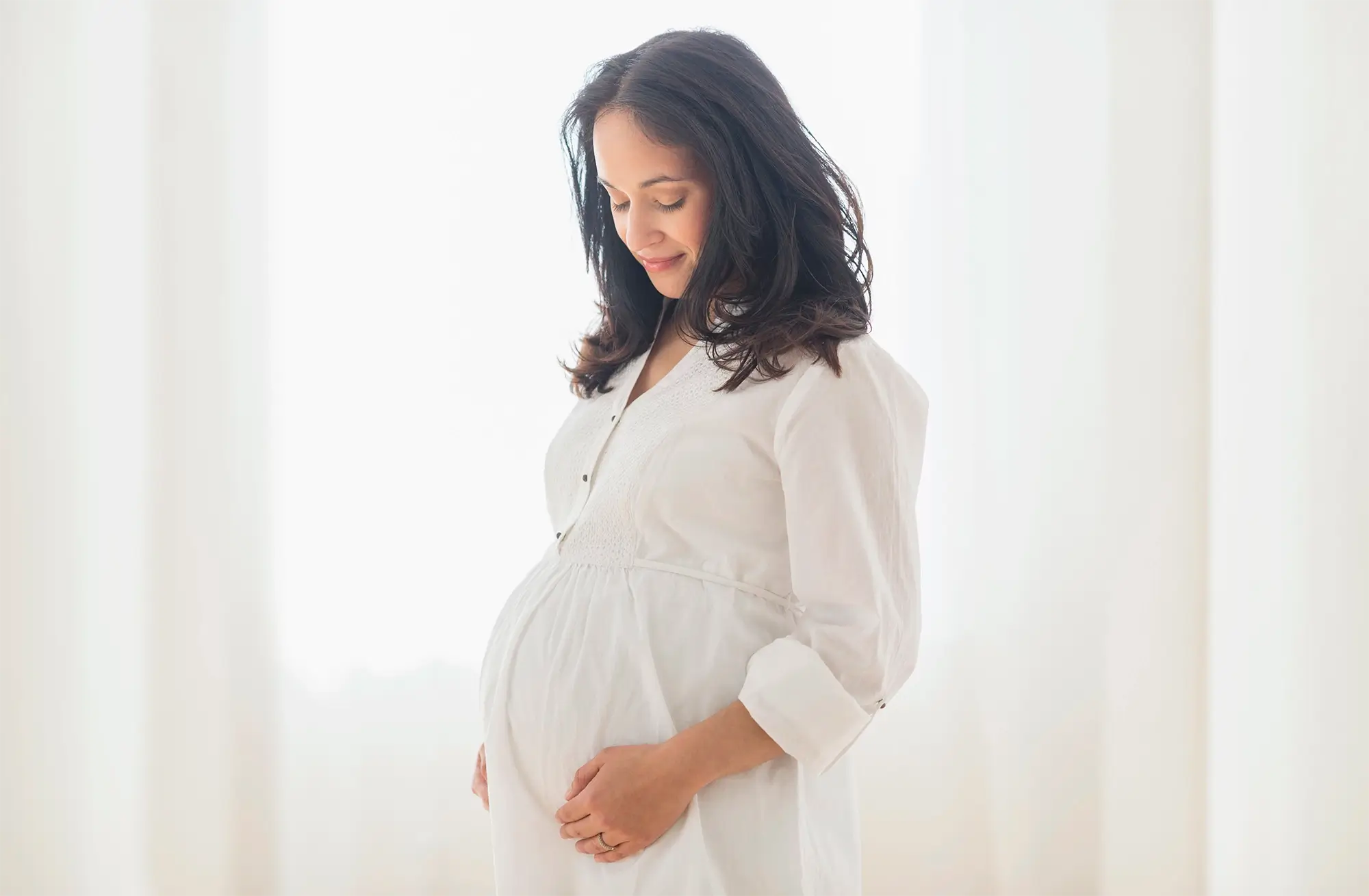 Expectant parent in a white dress holding their belly and smiling.